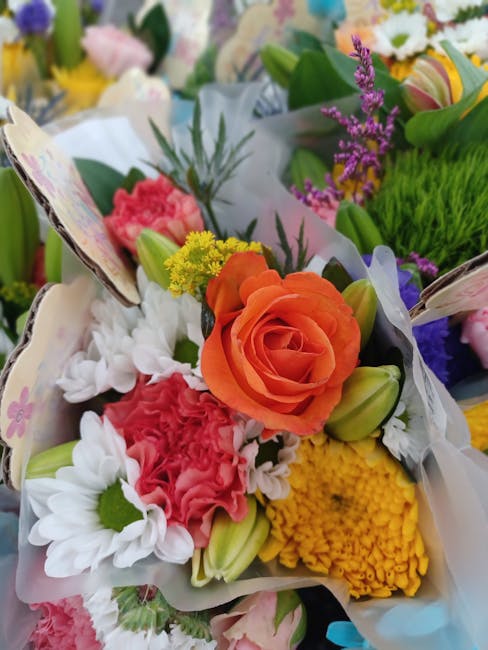 A vibrant floral arrangement featuring an orange rose at the center, surrounded by white daisies with green centers, pink carnations, yellow button chrysanthemums, and purple statice flowers. The bouq