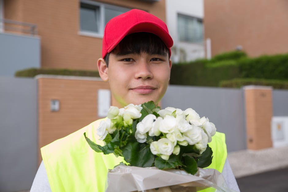 A young delivery person wearing a red cap and a high-visibility yellow vest is holding a bouquet of fresh white roses wrapped in white tissue paper, standing outdoors in a residential area with modern