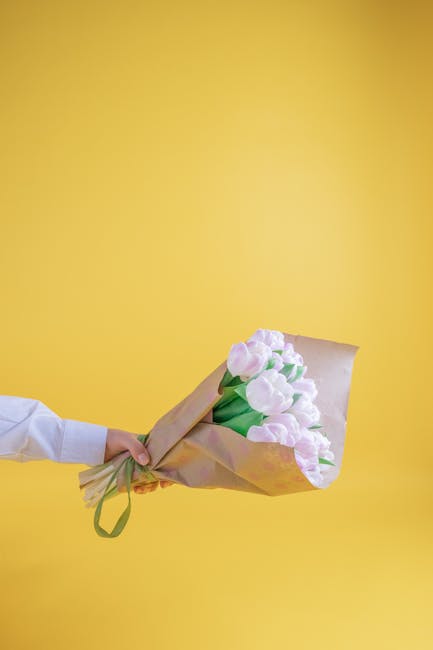 A person's hand, dressed in a white shirt sleeve, extends from the left side of the image holding a bouquet of fresh, pale pink tulips wrapped in brown kraft paper and secured with a green ribbon. The