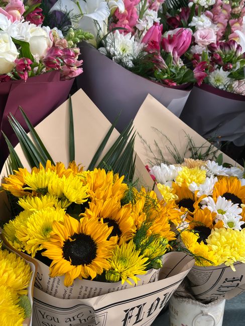 A vibrant display of fresh floral bouquets, featuring a collection of sunflowers with bright yellow petals and dark centers, arranged in white wrapping paper that mimics newspaper print. Behind them,