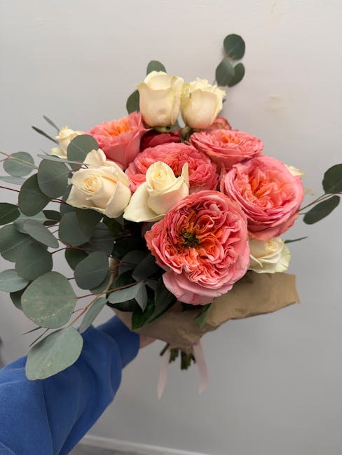 A bouquet of fresh flowers featuring large, ruffled pink ranunculus and creamy white roses, accented with rounds of eucalyptus leaves, wrapped in brown craft paper. The arrangement is held by a person