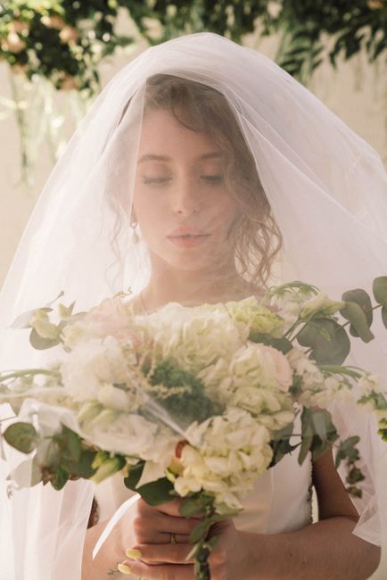 A bride holding a floral bouquet in front of her with a gentle, serene expression, dressed in a wedding gown and veil. The bouquet features a mix of white and blush pink roses, hydrangeas, and greener