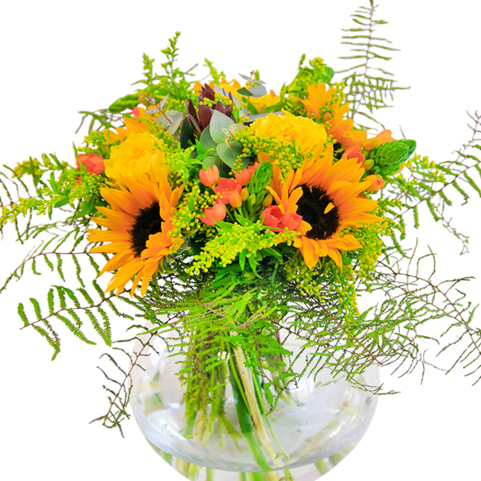 The sunlight-infused beauty of this bouquet captures the exuberant energy of a perfect New Southgate day. Bursting from a crystal-clear, rounded vase, towering sunflowers and velvety yellow roses intertwine, their petals fanned open as if reaching for the skies above nearby Bethune Park. Each sunflower boasts a rich, brown heart framed by a corona of golden-orange rays, while the garden roses offer a softer, spiral texture-luxurious in their fullness. Moments of vivid red, brought by clusters of hypericum berries, twinkle amid the greenery like cheerful lanterns lighting up the lush undergrowth. Fern leaves arch elegantly around the arrangement, adding gentle movement and depth, reminiscent of wild, rambling gardens seen along Green Road. Sprigs of fine filler, hinting at goldenrod, introduce clouds of miniature yellow blooms, making the entire display feel as fresh as a morning after rain. Eucalyptus leaves, subtle in their blue-green hue, weave among the larger leaves, adding a cool, fragrant counterpoint. The stems rest in crystal water, every detail visible through the vase, emphasizing the arrangement's honest craftsmanship. This bouquet is a sensory delight-delicate fragrances mingle with the velvety feel of rose and sunflower, while the lively colours lift the heart. Whether celebrating a birthday, welcoming someone home, or simply brightening a room, this arrangement is a vibrant testament to the artistry and warmth of New Southgate floristry.
